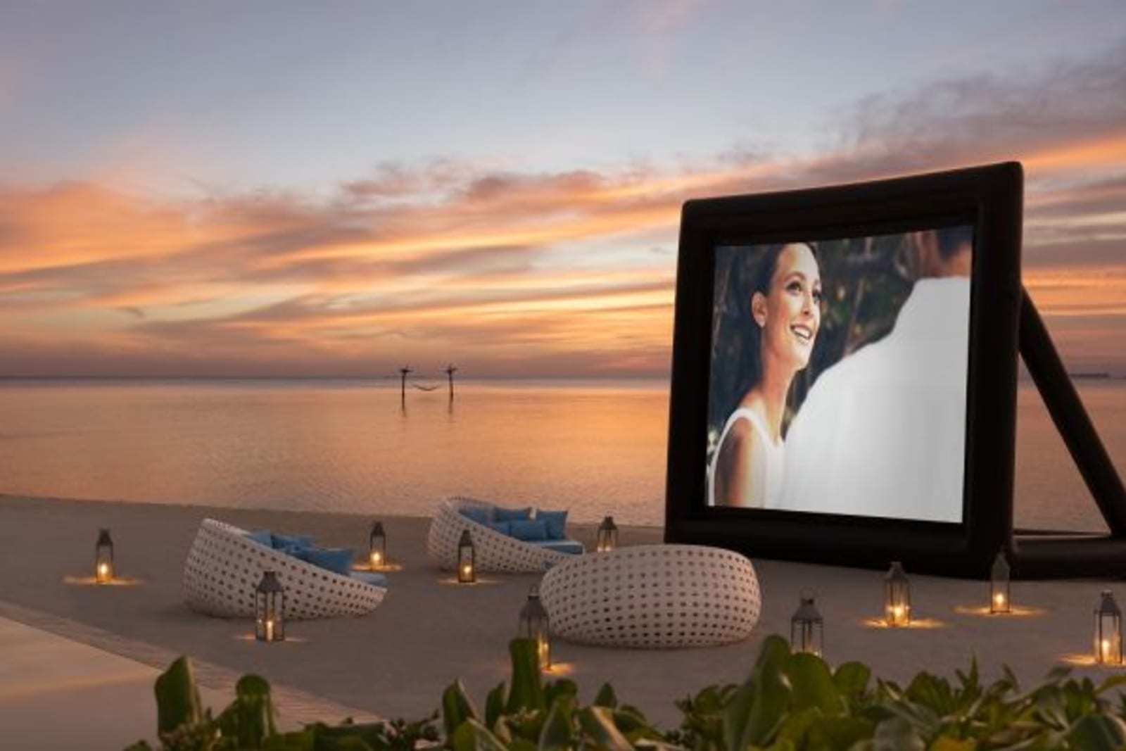 Large projected movie screen on beach front with large cushioned chairs at sunset