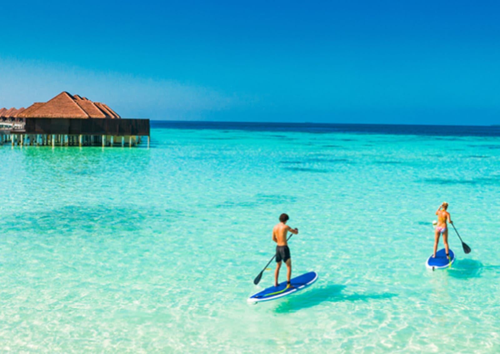 Two people on stand-up paddleboards on a clear ocean, with rows of bungalows in the background