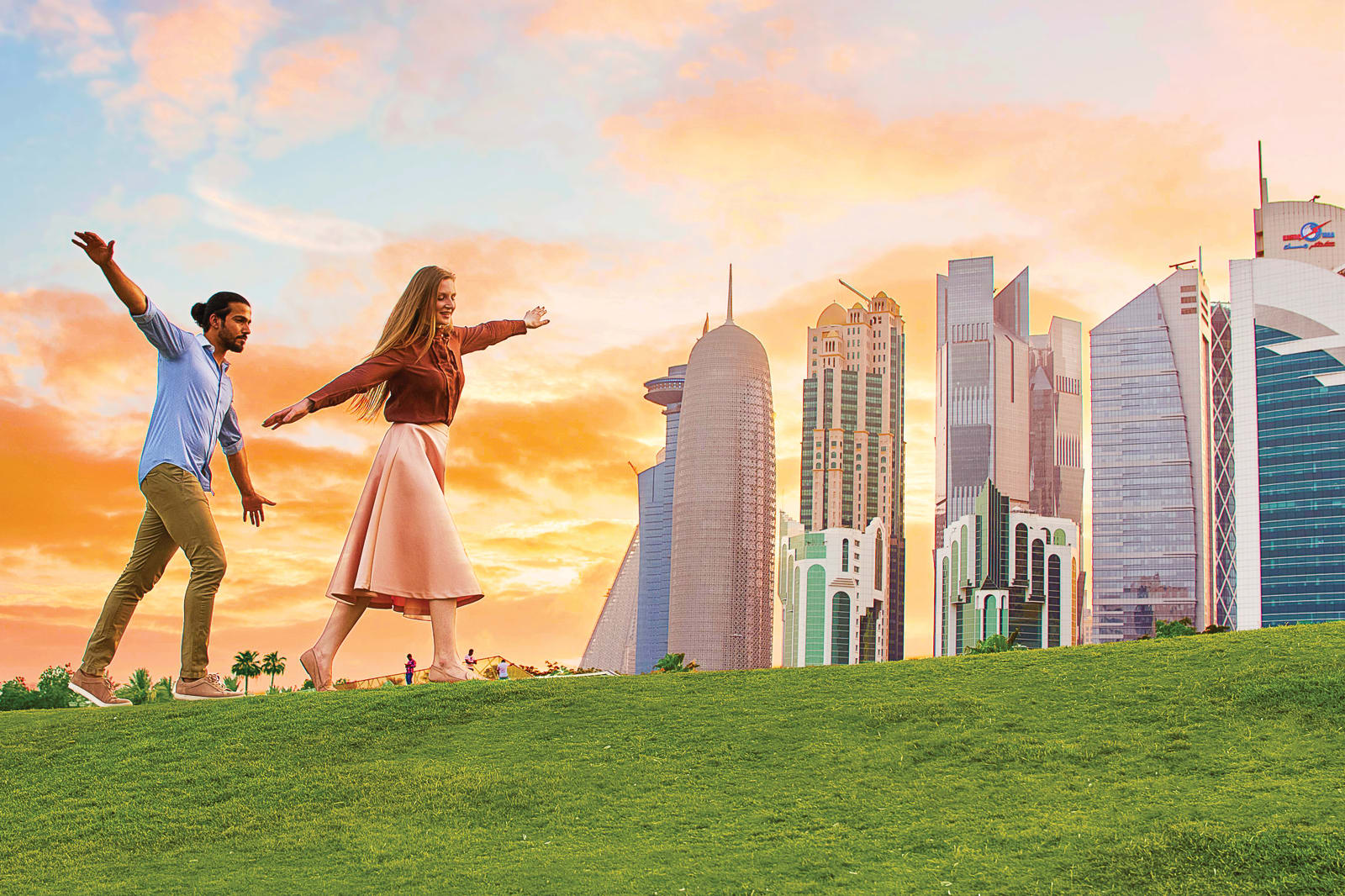 Two people with their arms out walking along grass at sunset with the Qatar city skyline in the background