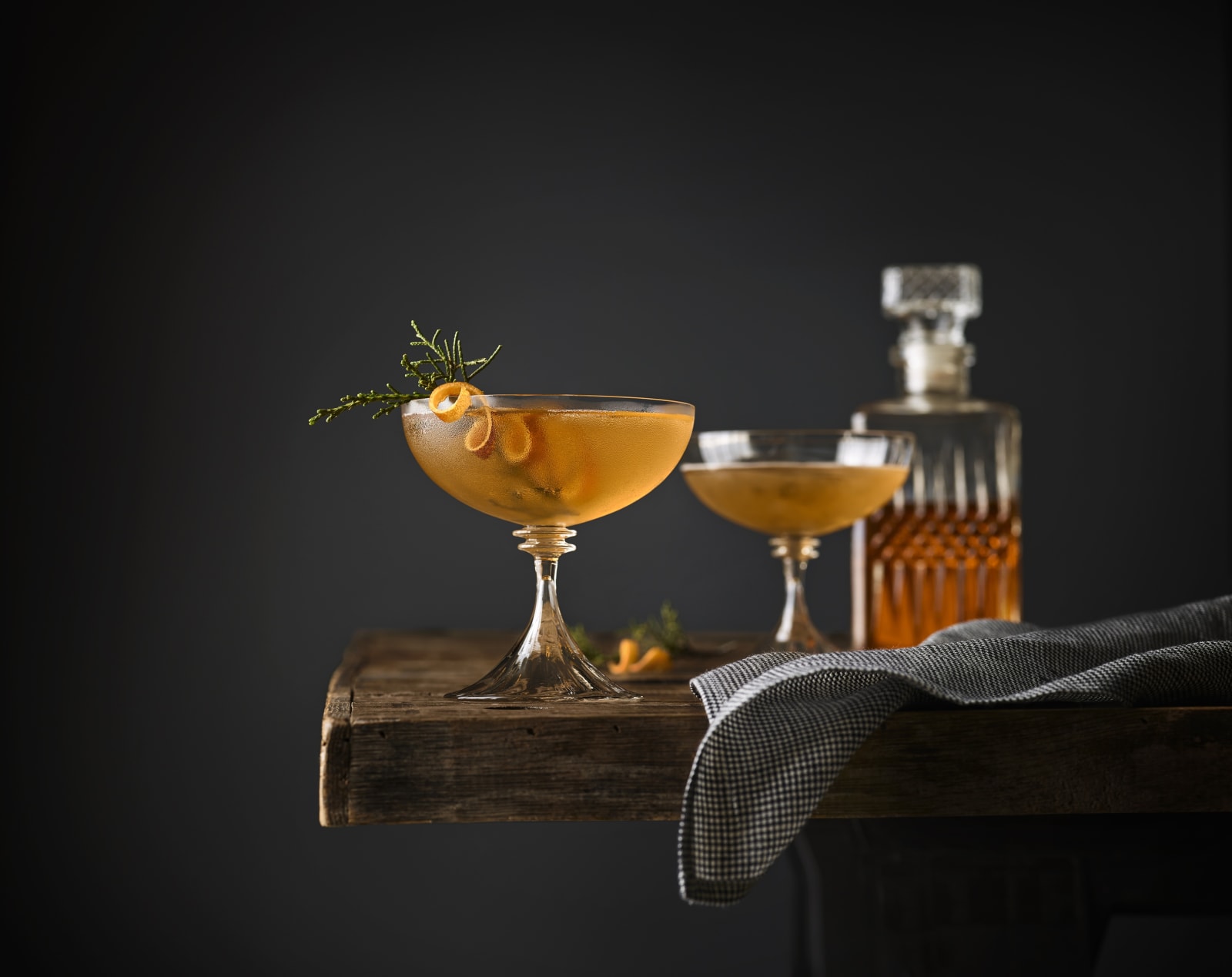 Still life of two Manhattan cocktails garnished with Juniper on a table with whiskey decanter in the background.