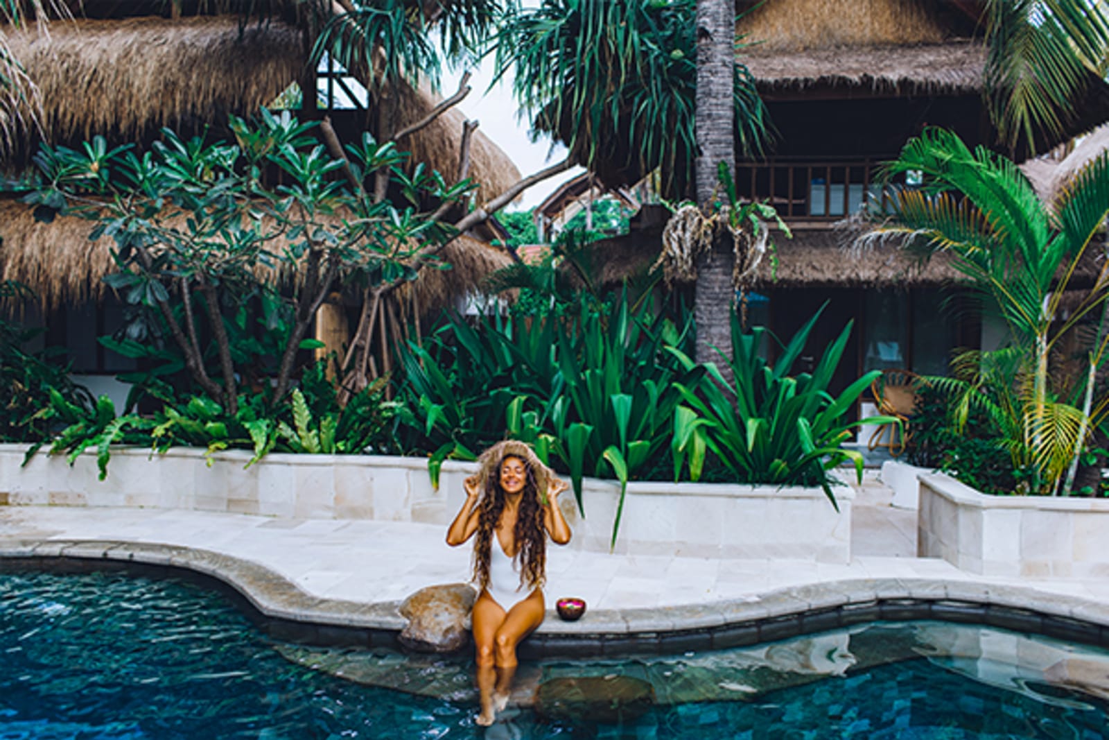 Woman in white swimsuit relaxing in a pool surrounded by lush, green plants and thatched roofs
