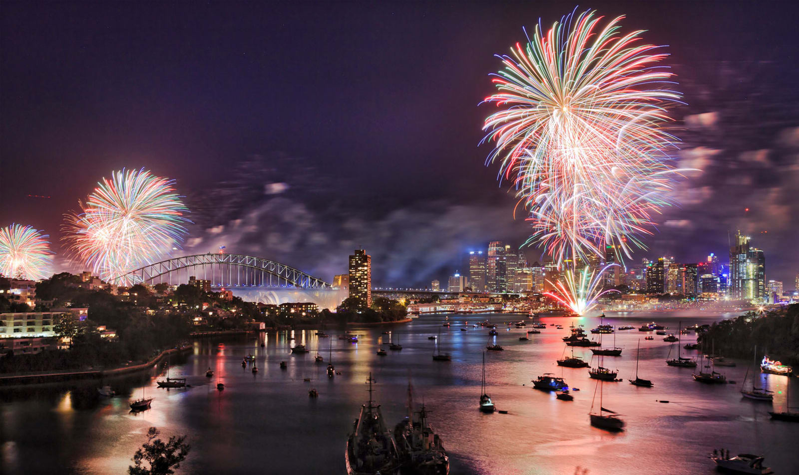 Sydney New Year eve fireworks over Harbour with bridge and city CBD buildings reflecting colourful fire balls in blurred water.
