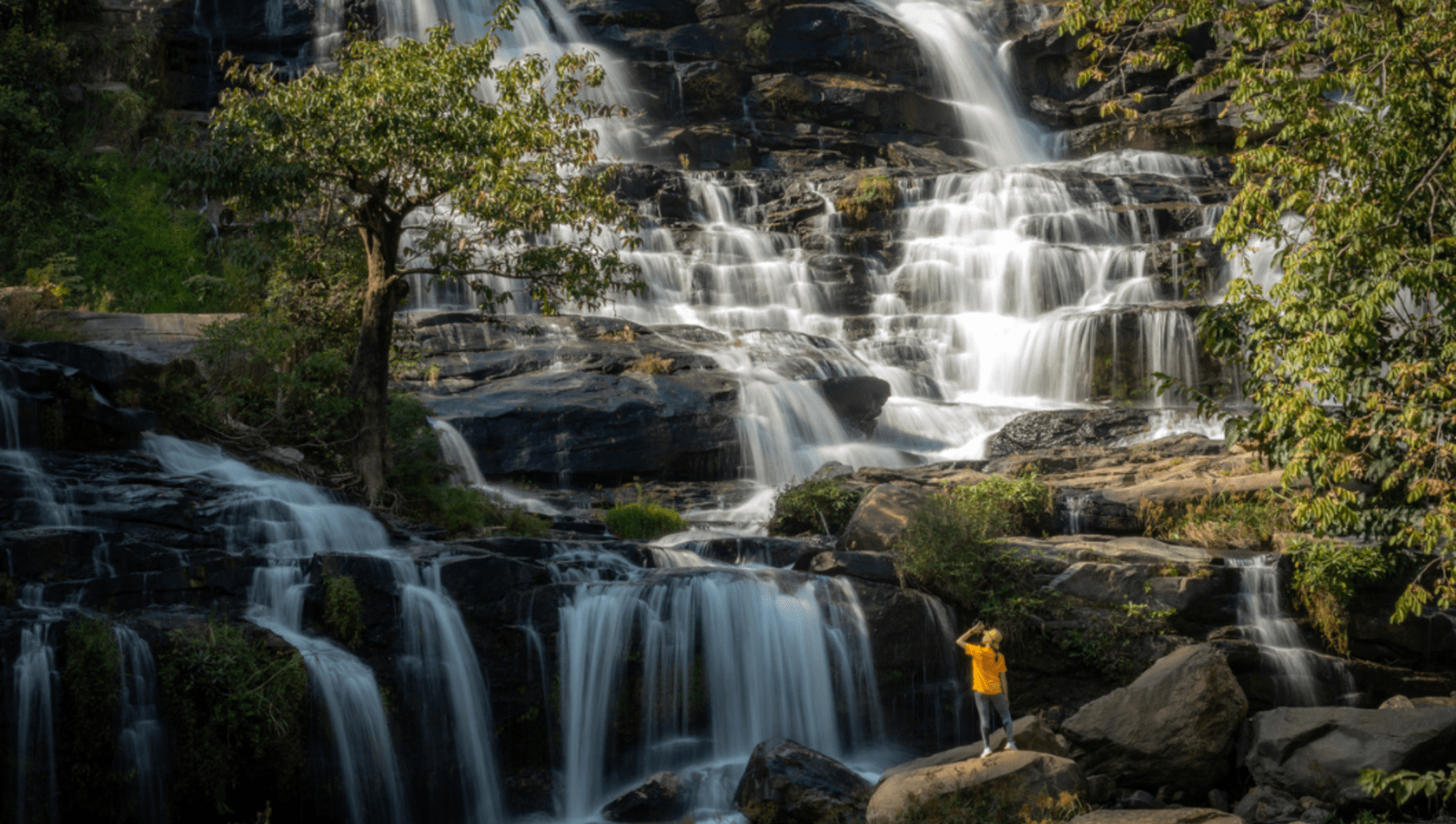 Lady in yellow jacket looking up at a big waterfall in chiang mai thailand