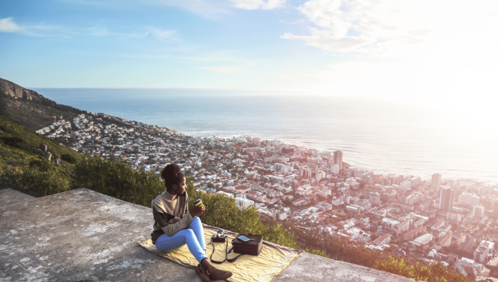 Lady sitting on yellow blanket while on top of a hill overlooking a city and the sea in capetown