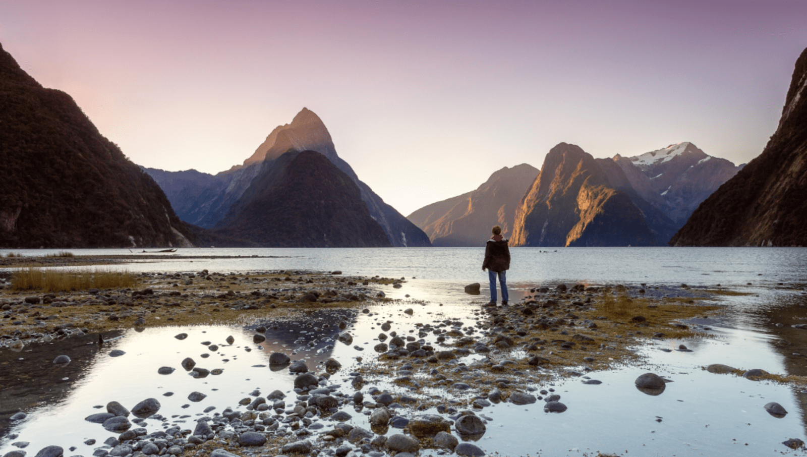 Person standing on rocks on the shore of a lake, looking at mountains across the lake