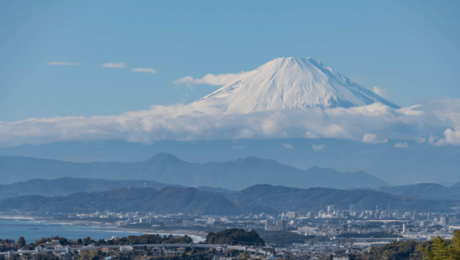 View of Mount Fuji covered in snow and surrounded by clouds from coastal town of Kamakura