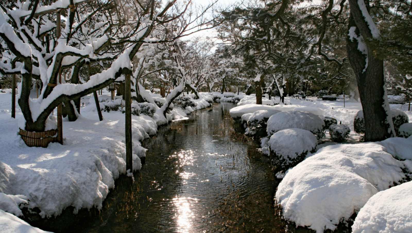 Small river in a garden surrounded by trees and ground covered in snow