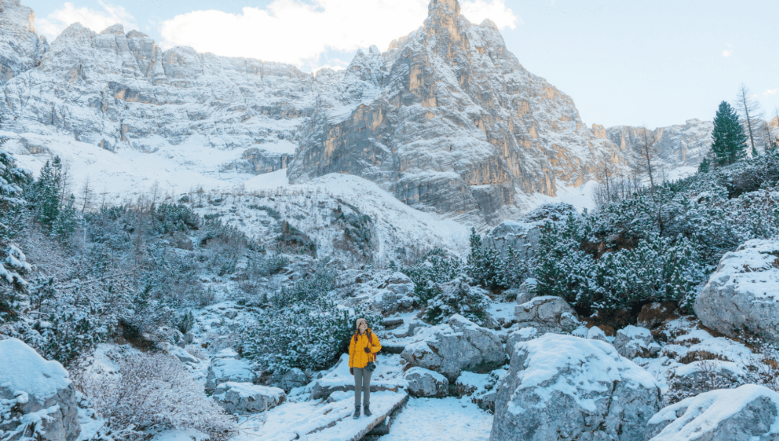Lady in yellow puffer jacket hiking in the Dolomites surrounded by snowy mountains and trees