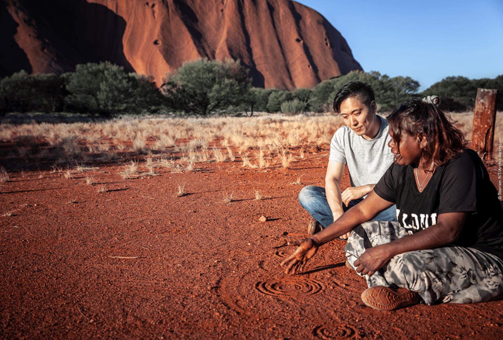 An Aboriginal woman traces stories in the sand for an onlooker beside Uluru