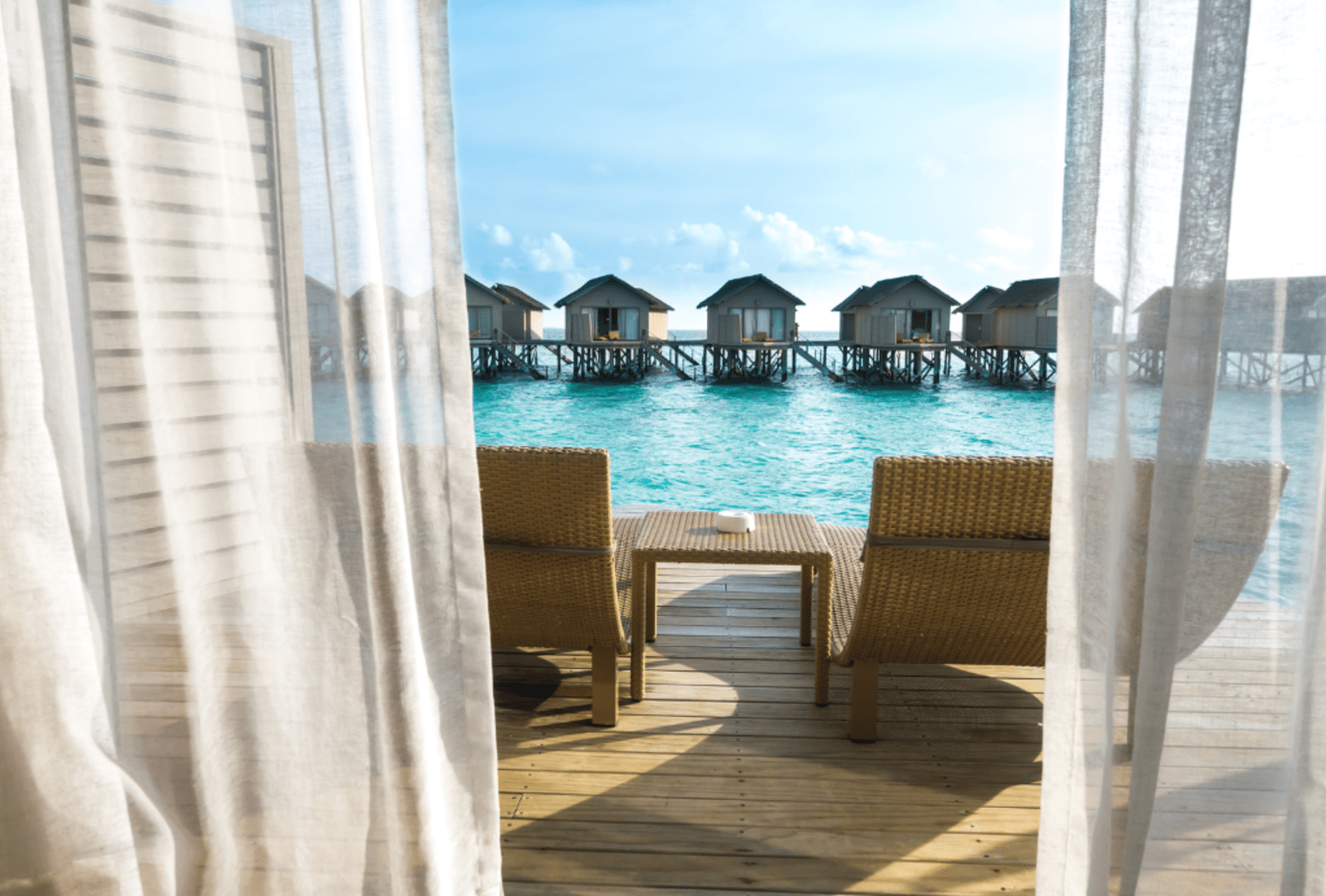 A view through thin curtains to a balcony overlooking calm, turquoise waters with arow of other overwater bungalows in the background