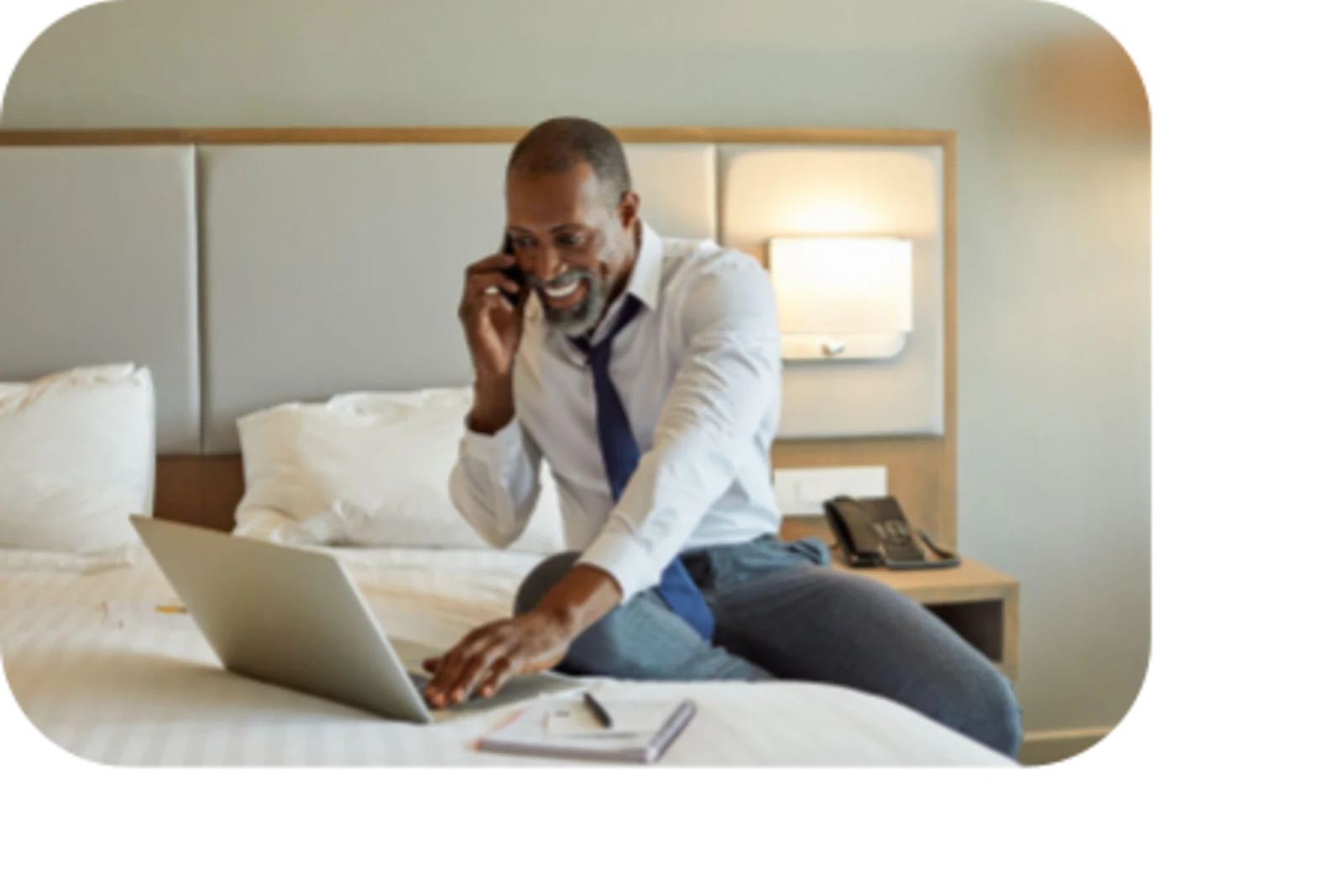 man seating on hotel room bed, using laptop and talking in phone