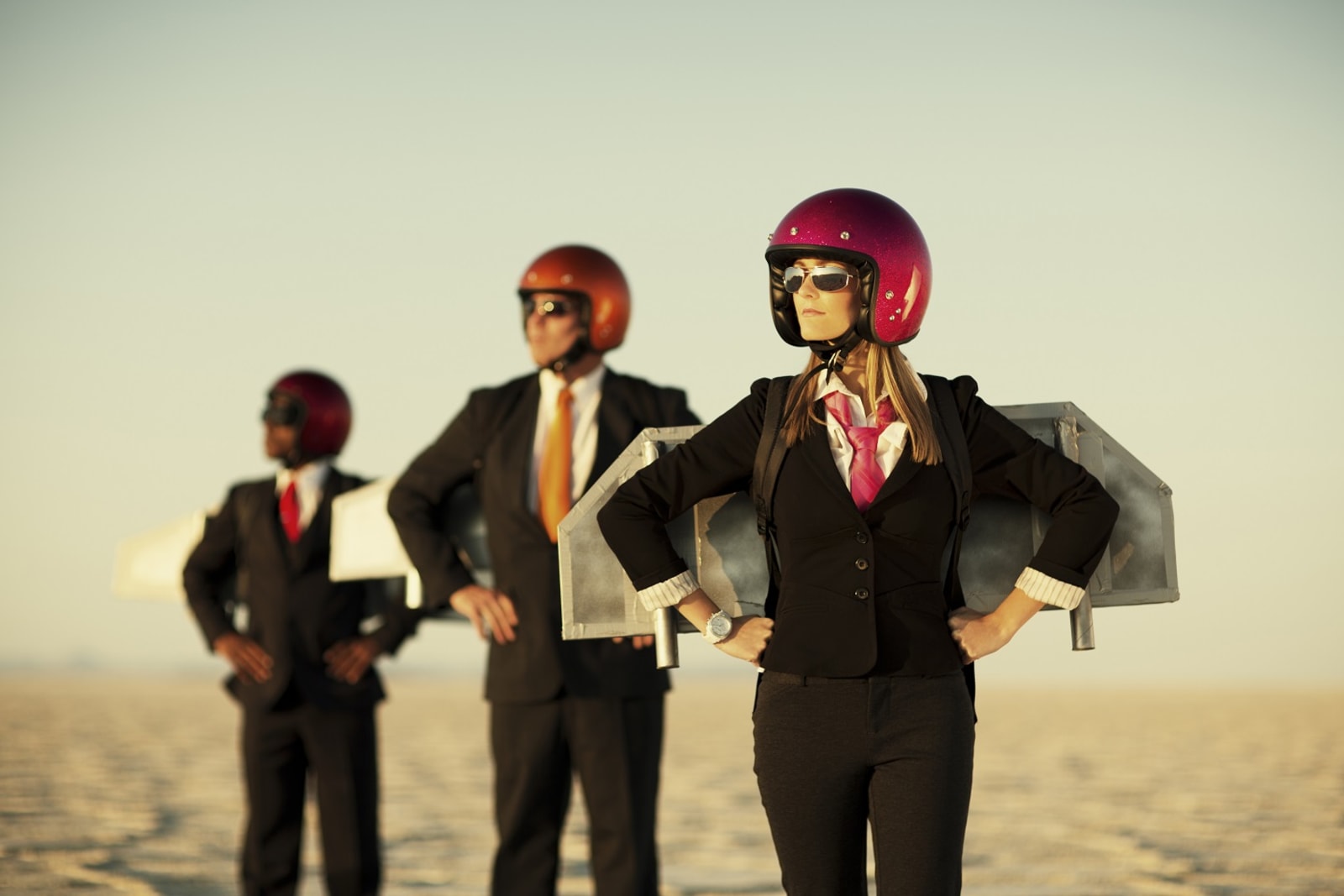 3 people in business suits wearing red crash helmets and costume space wings, in a desert