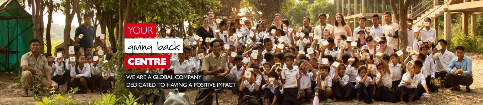 Your giving back centre - we are a global company dedicated to having a positive impact. Group of multi-racial students and teachers holding papers in the air framed by trees and a school building