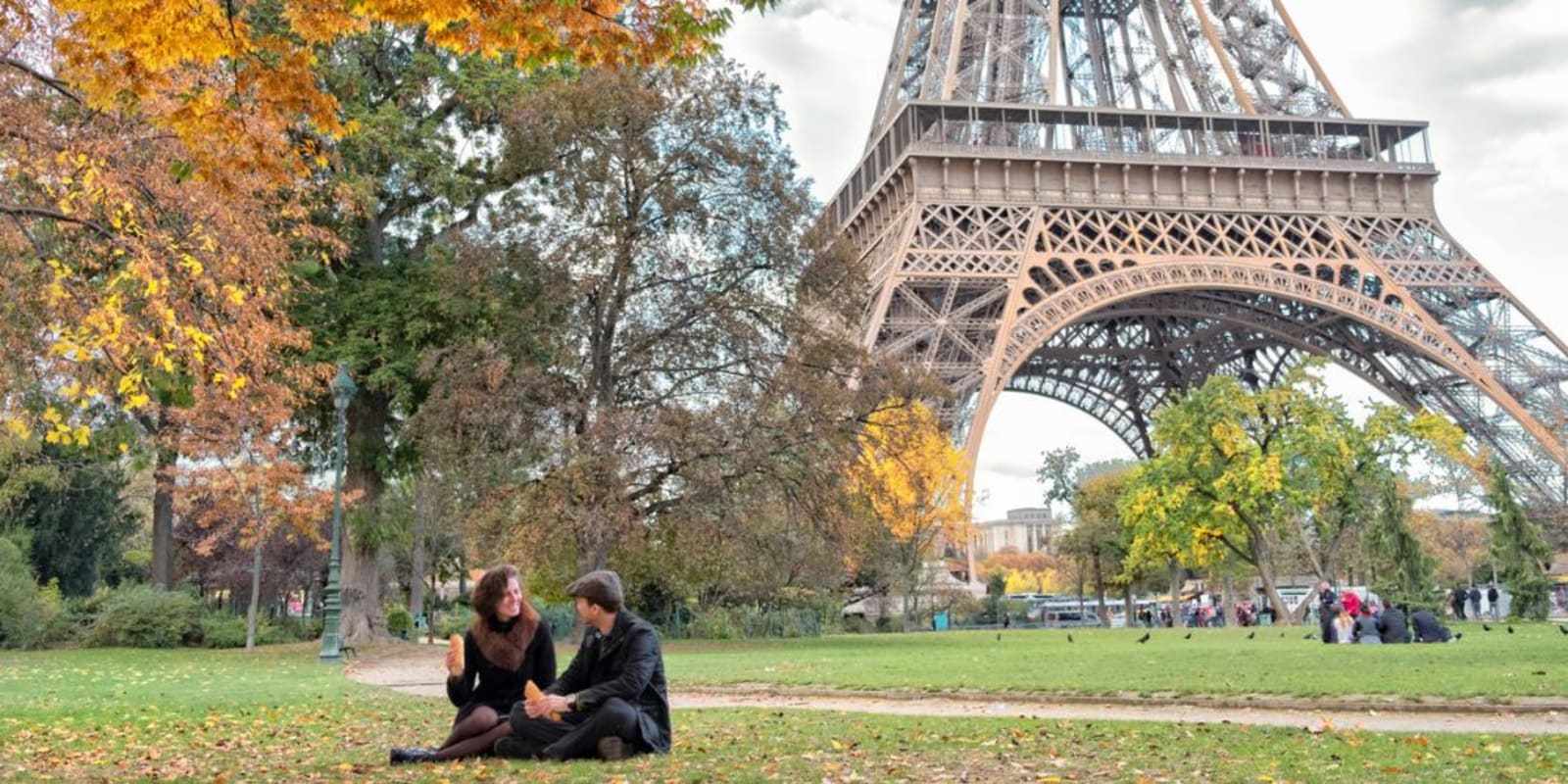 A couple sitting on the grass in front of the Eiffel tower eating a Baguette