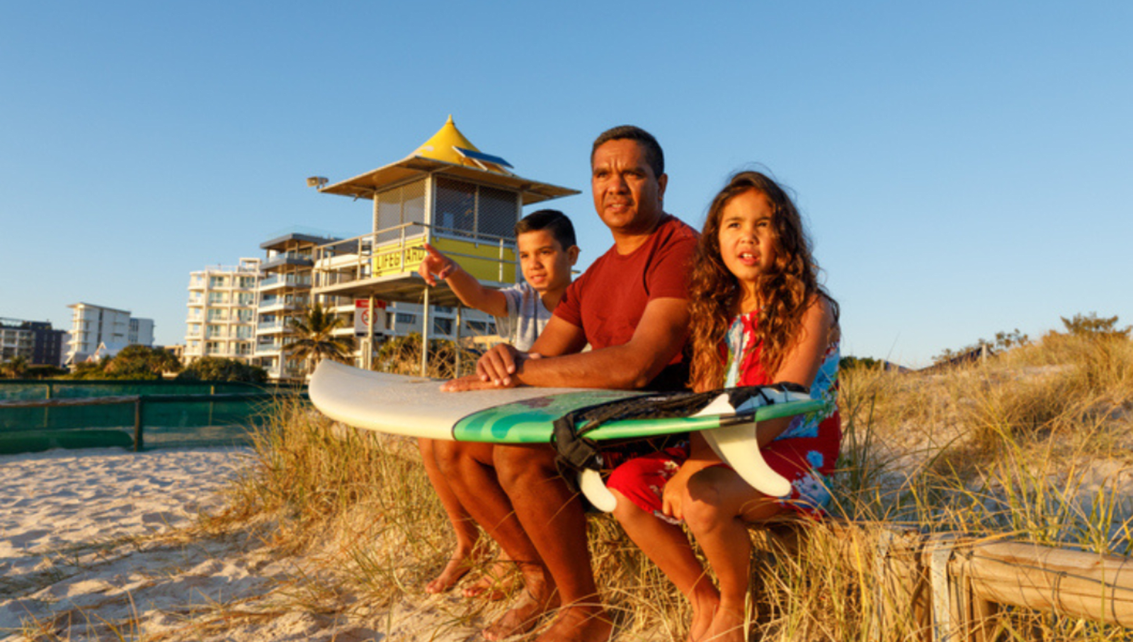 Two children and adult sit near lifesaving tower on Gold Coast Beach with surfboard across their laps