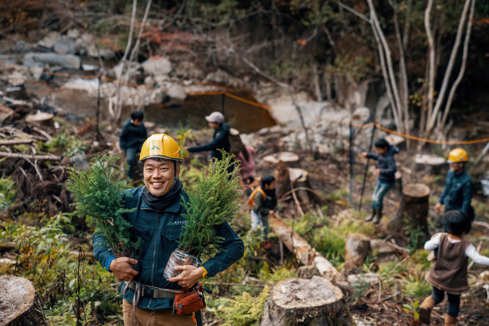 Man holding two small trees smiling to camera while other people plant trees in background