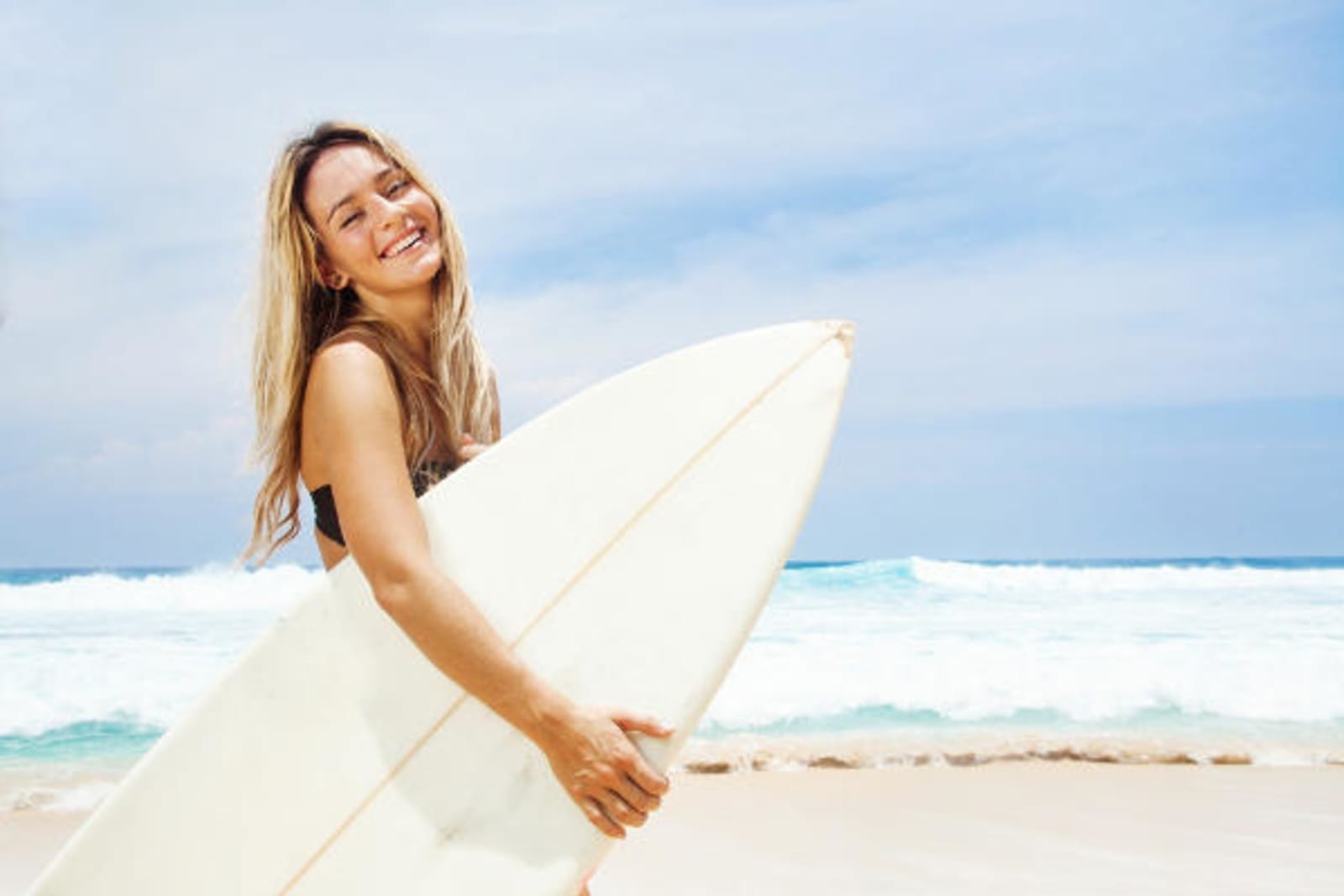 Woman smiling on a brightly lit beach, holding a white surfboard