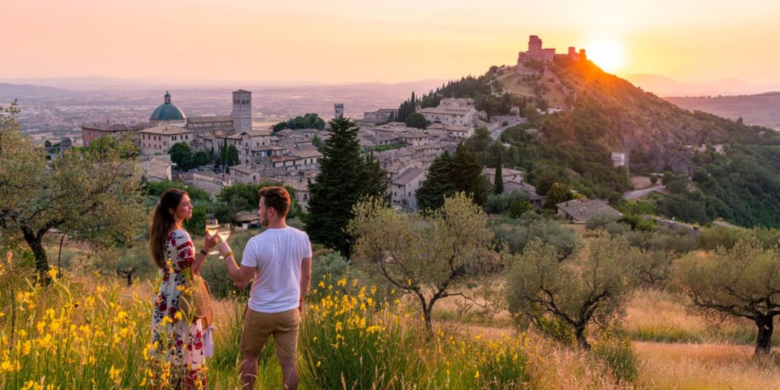 A couple drinking a glass of wine together on a hillside as the sun sets in the background