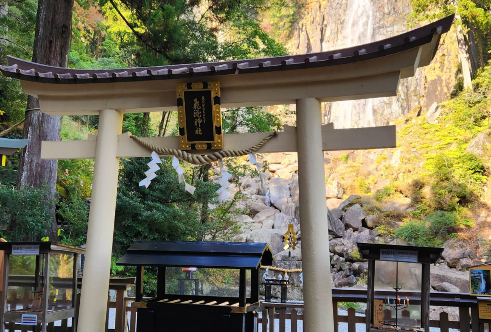 A traditional torii gate stands before a tall waterfall drenched in sunlight