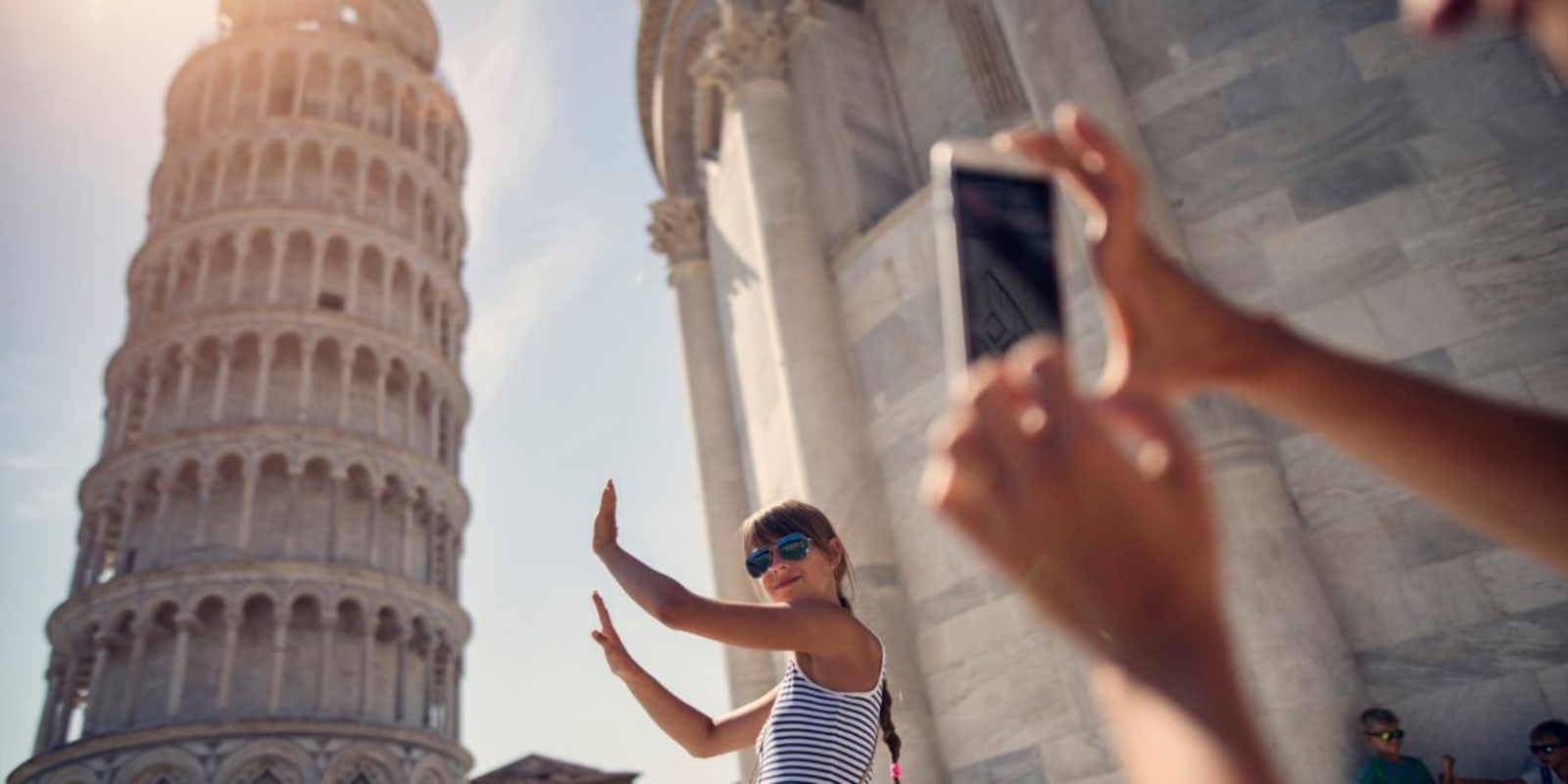 A person using their phone to take a photo of someone pretending to hold up the leaning tower of Pisa