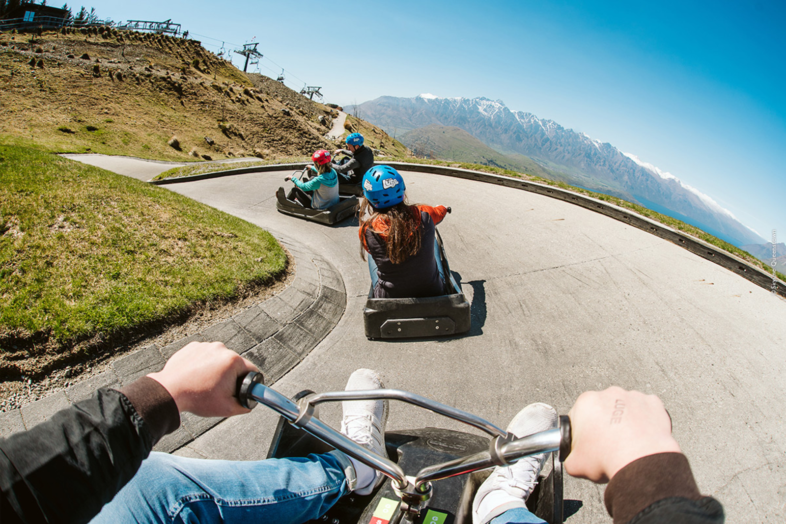 A family rides luges down a steep course with The Remarkables mountain range in the background, Queenstown NZ.