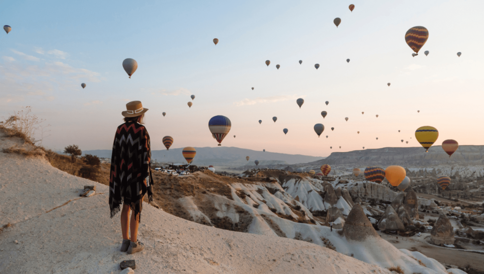lady standing on cliff looking at lots of hot air balloons in the sky