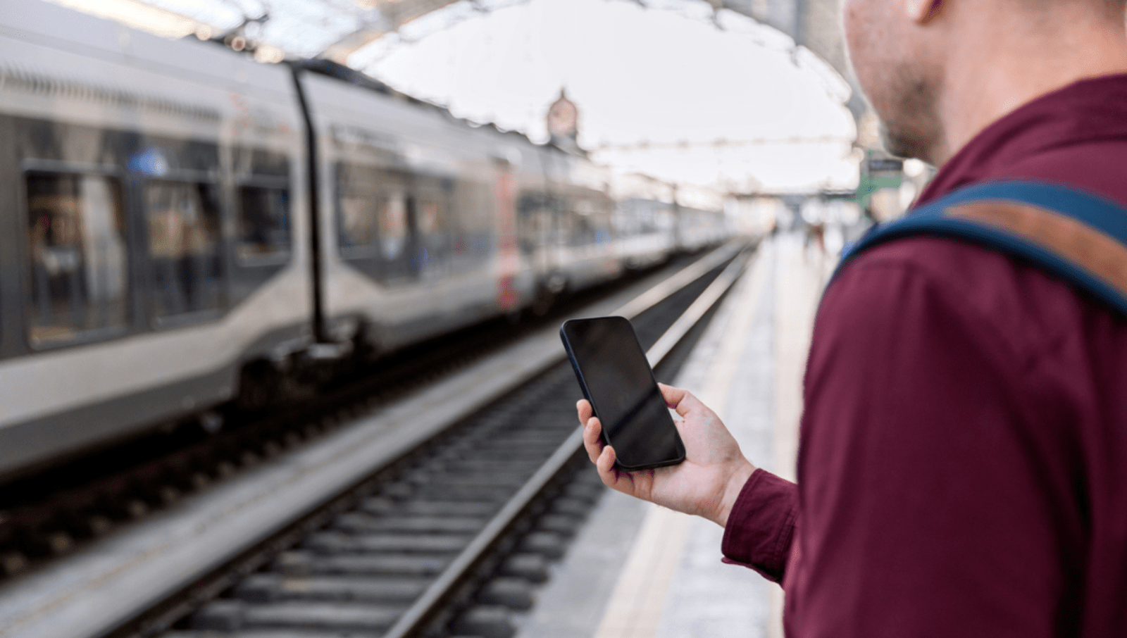 Man wearing red shirt holding phone out in front of him at a train station as a train passes by