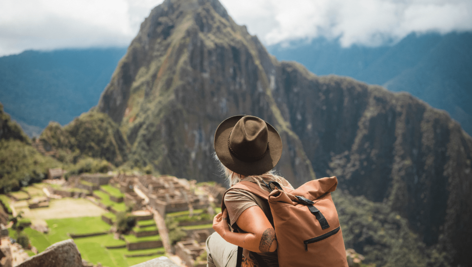 Tourist with hat and backpack standing on sliff overlooking maccu picchu