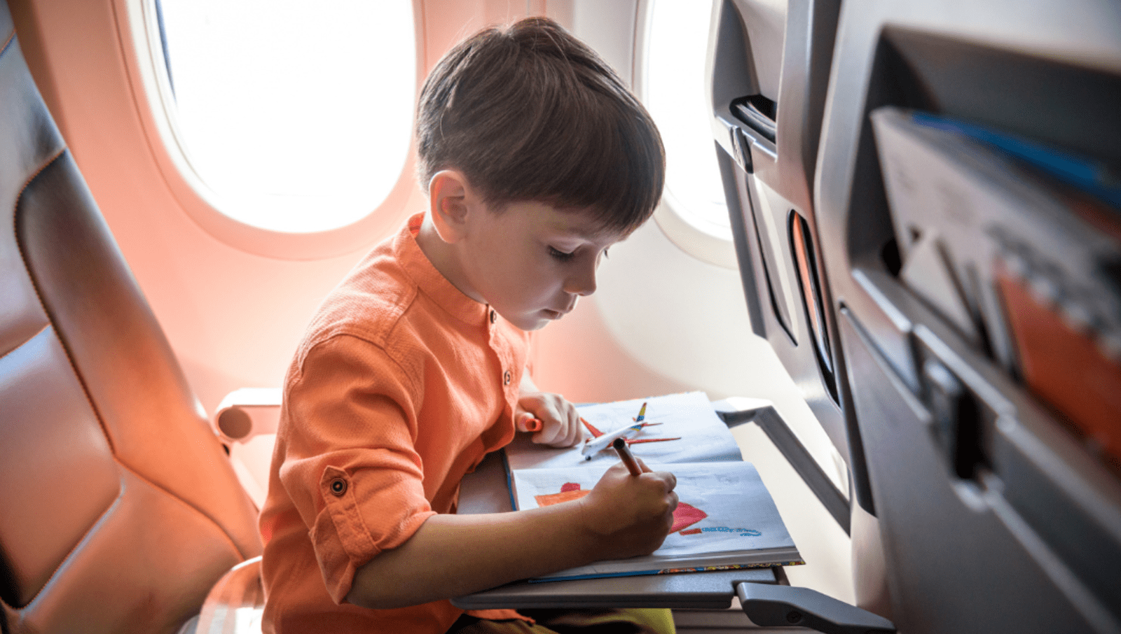 Child in orange shirt drawing with crayons on pen with toy airplane sitting on drawing book 