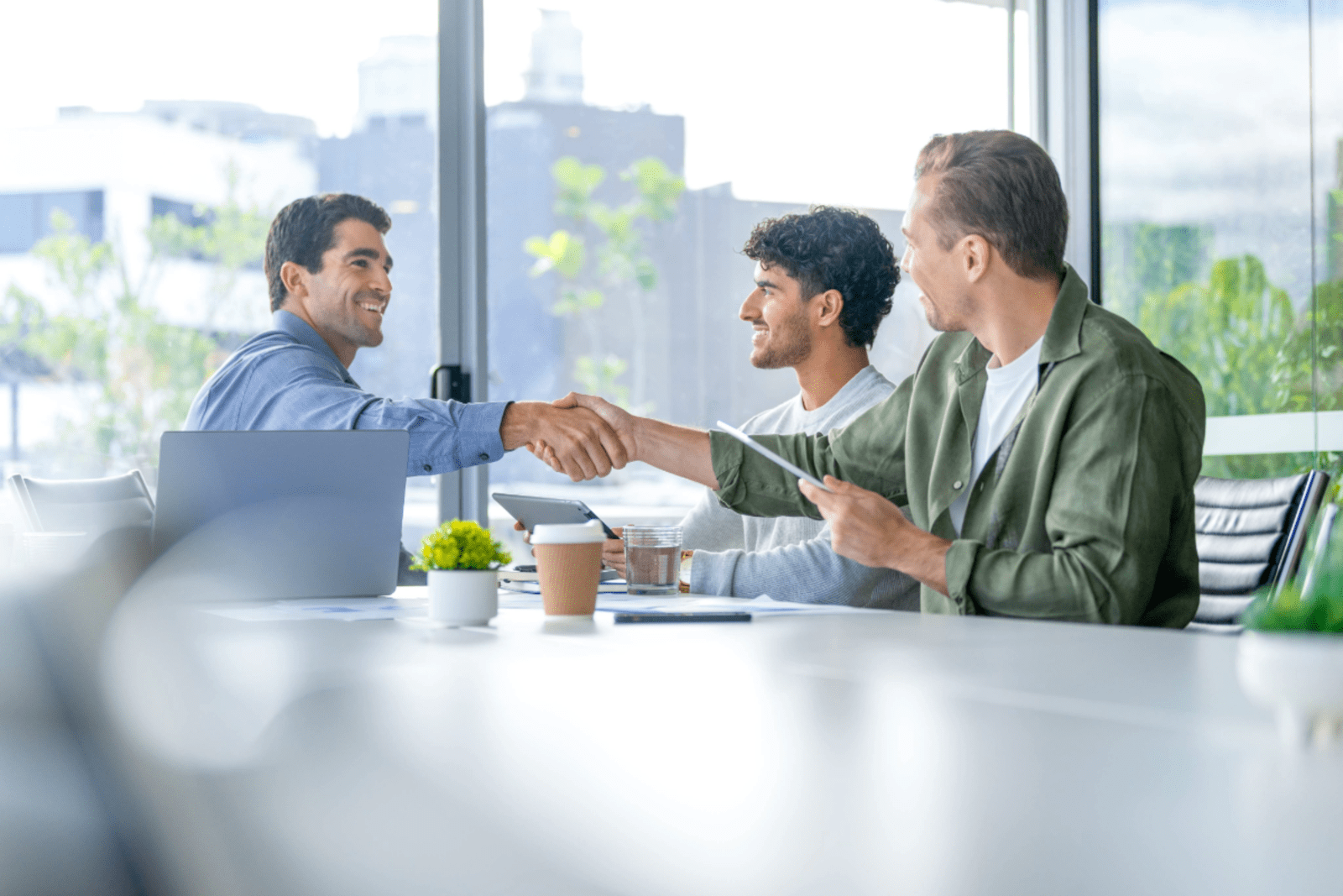 three business men sitting around table in board room with two of them smiling and shaking hands
