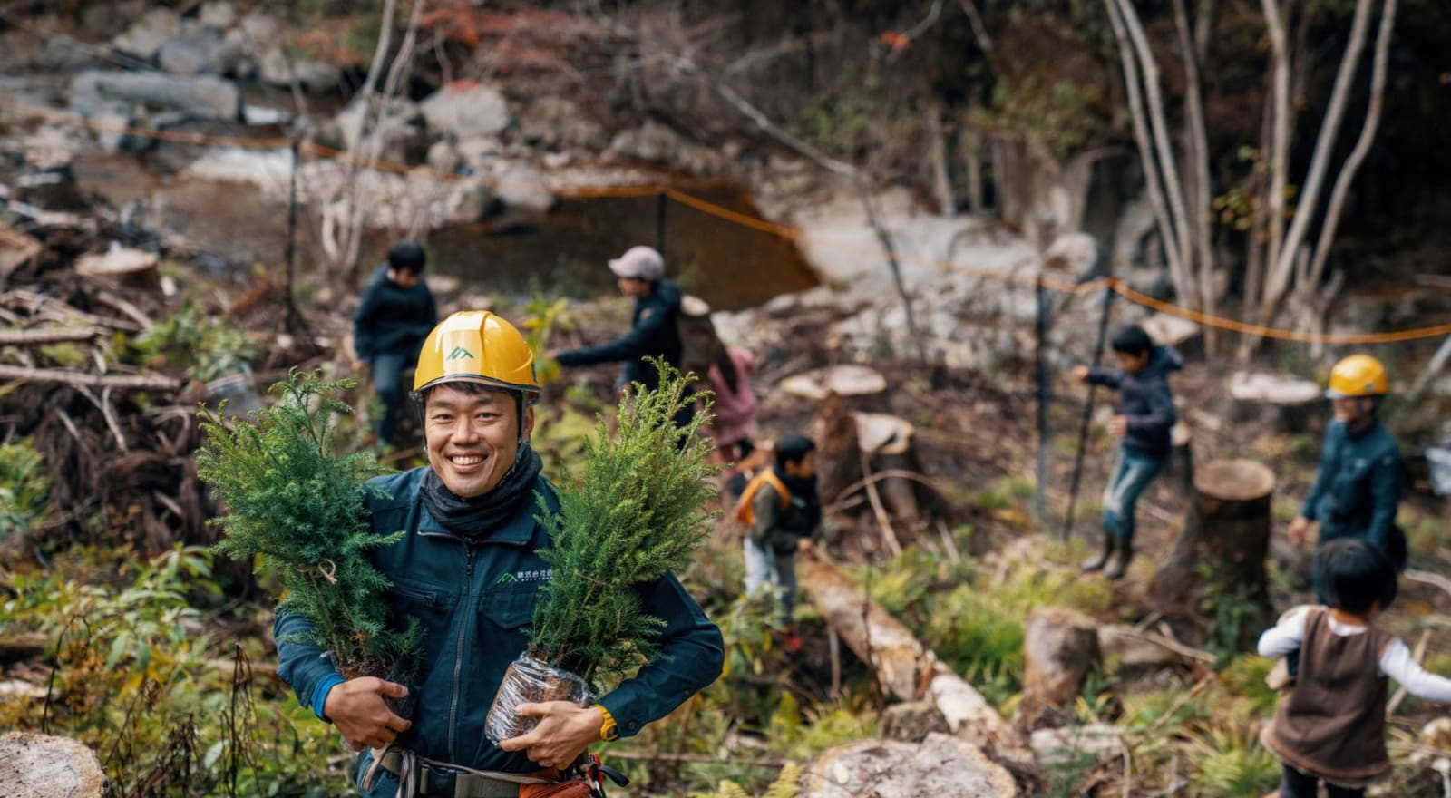 Man holding two small trees smiling to camera while other people plant trees in background
