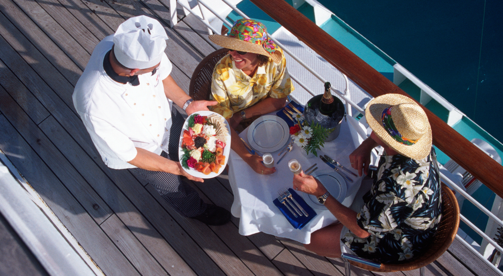 chef serving plate of food to couple sitting at table on outside deck of cruise ship