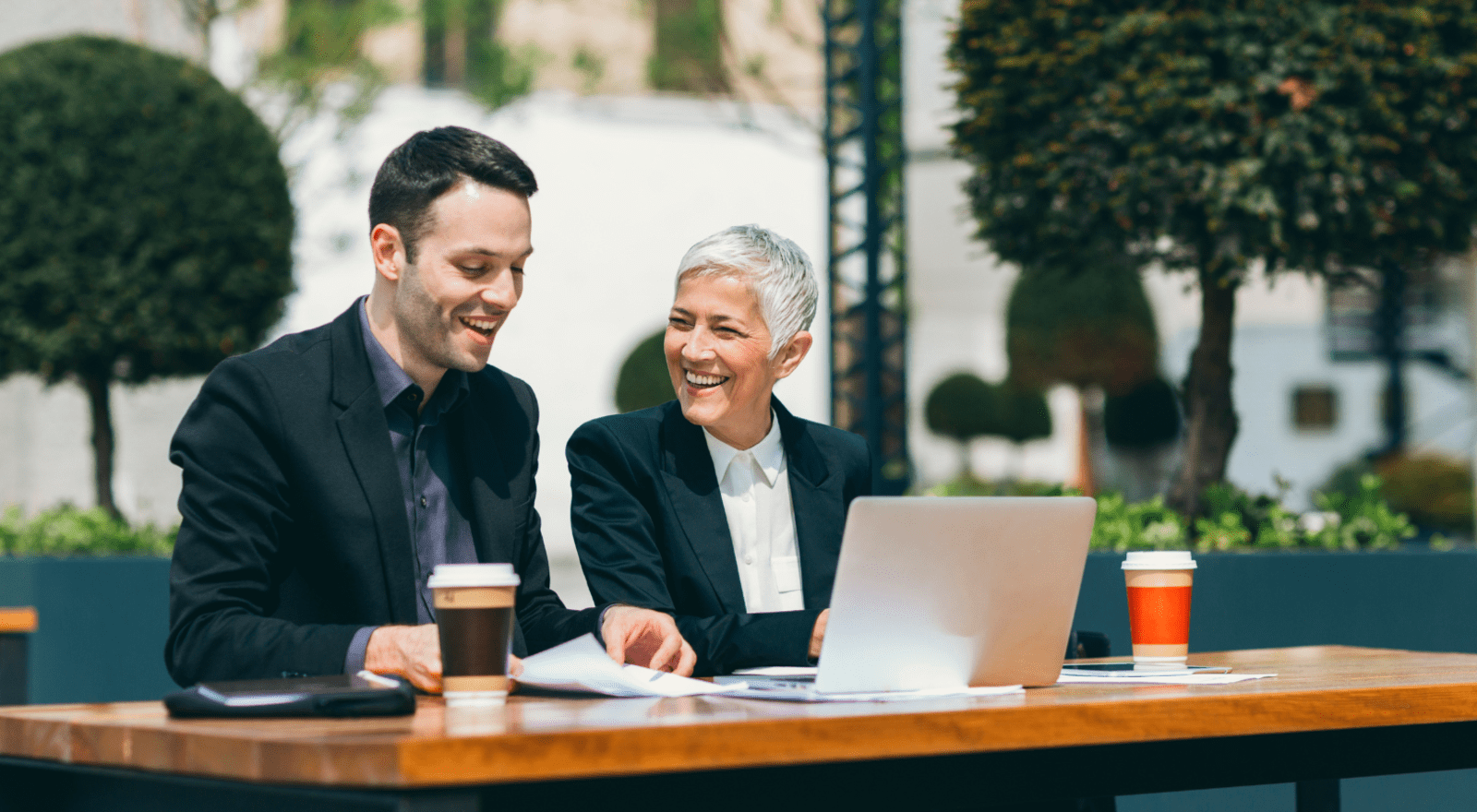 two business people sitting at a park beck smiling and laughing with a laptop and coffees
