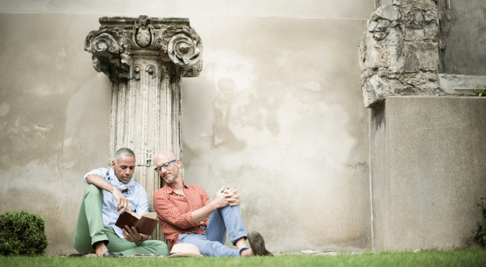 two men sitting on grass in front of stone columns reading a book