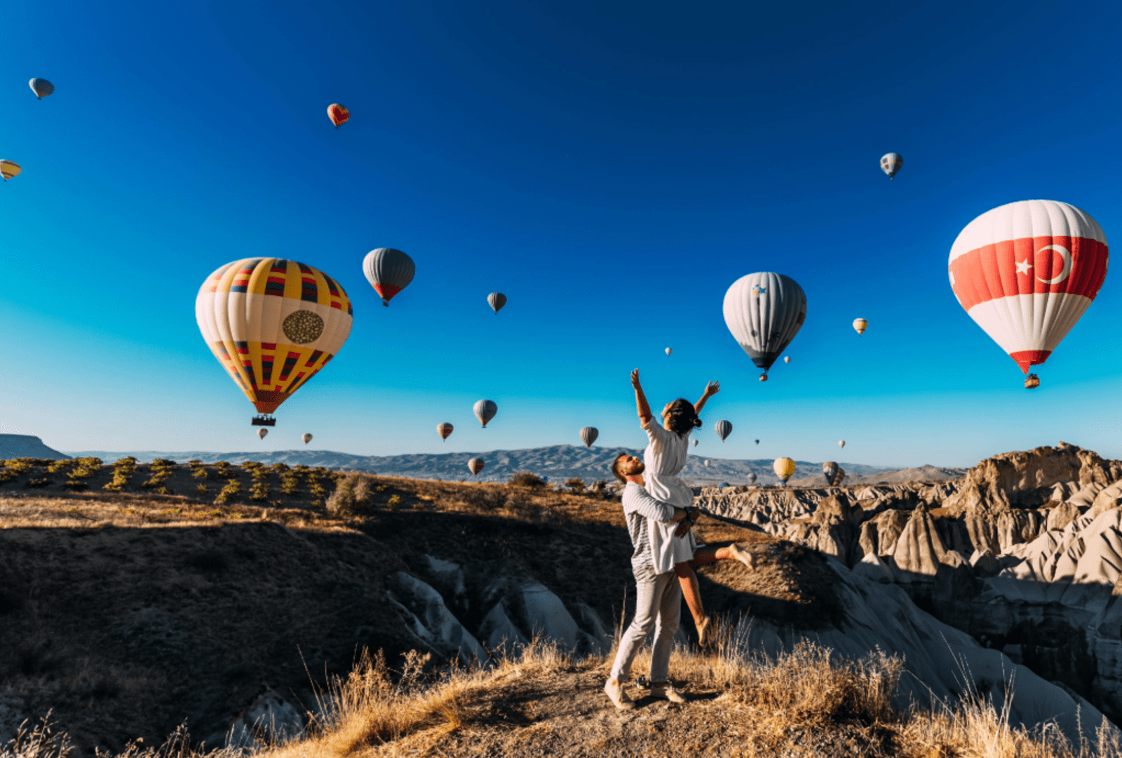A man picks up a woman under a blue sky full of hot air balloons in Cappadocia, Turkiye