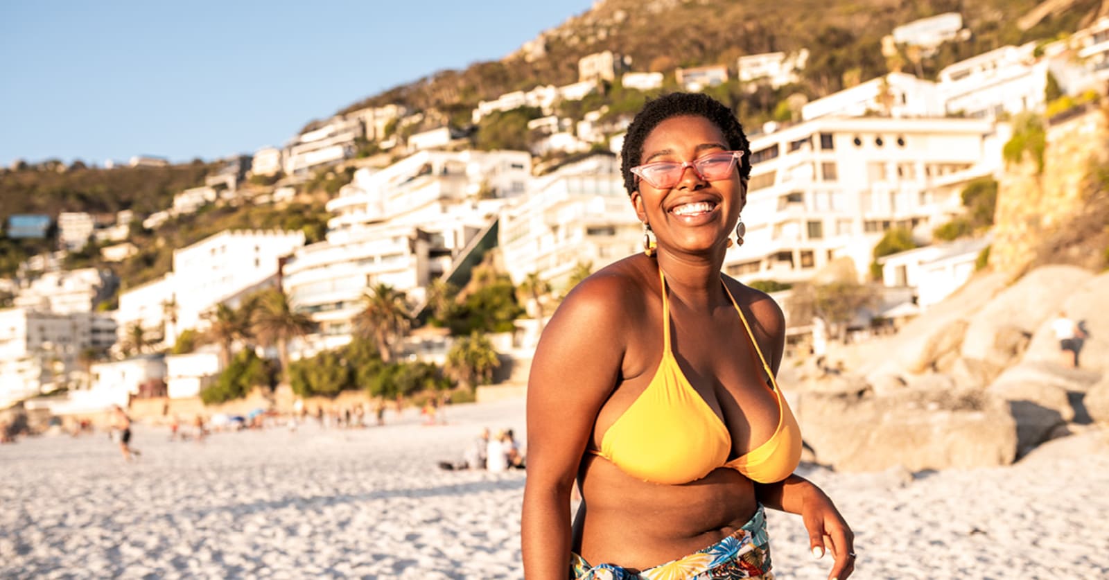 Woman with sunglasses and yellow bikini top smiles on a beach with bright hotel buildings in the background