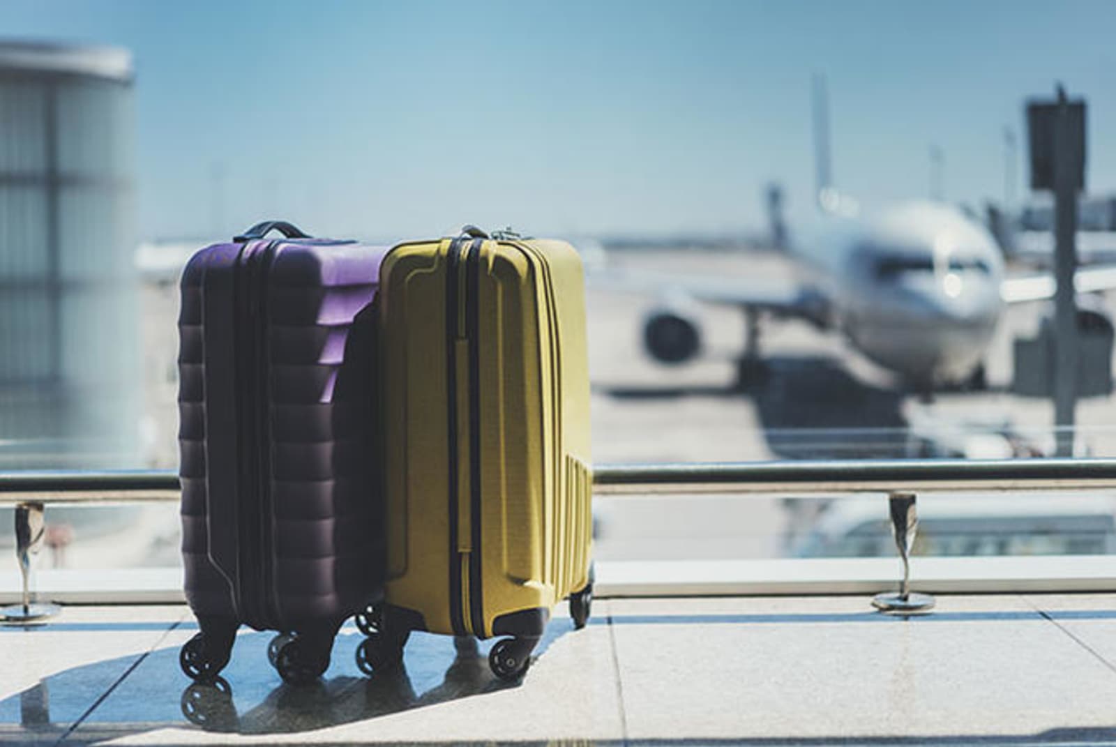 Two rolling luggage cases - one purple, one yellow - in an airport, in front of a large window looking out at an airplane