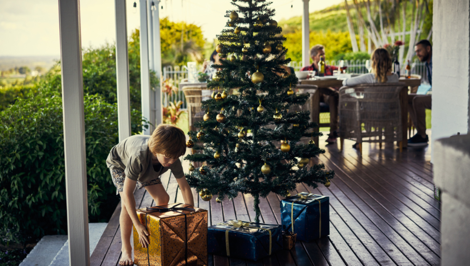 child picks up present wrapped in gold, sitting next to a green and blue present sitting under a christmas tree on a veranda. Family sittings at wooden table in background 