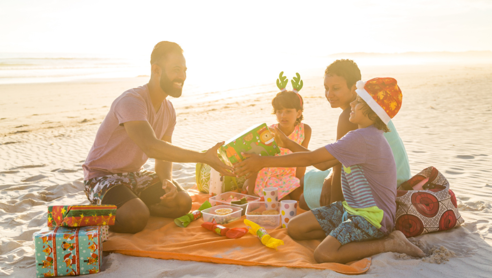 Family of two adults and two children exchanging Christmas presents on the beach with food and crackers in front of them