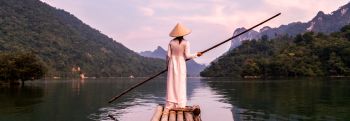 Woman wearing traditional Asian clothing holding a long wooden stick on a canoe floating on a river in between two mountains