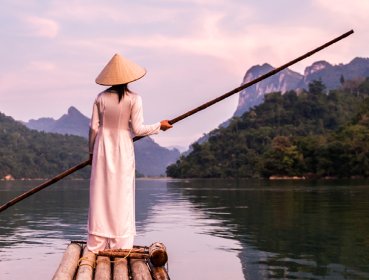 Woman wearing traditional Asian clothing holding a long wooden stick on a canoe floating on a river in between two mountains