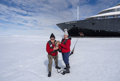 Couple bundled in red jackets holding whale bone on the ice beside the Scenic Eclipse II ship
