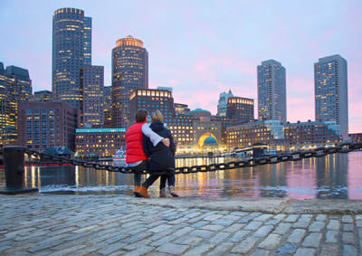 couple sitting on railing looking over at Boston across the river at early evening