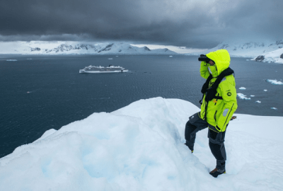 Expedition traveller in yellow jacket surveying icebergs and mountains from a snowy Antarctic ridge with the ship below