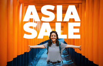 A smiling traveler stands with arms outstretched along a pathway of vibrant orange torii gates at a Japanese shrine, wearing a striped shirt and backpack. Bold white text at the top reads “ASIA SALE.”