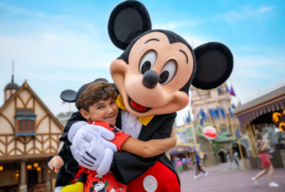 Young boy smiling and hugging Mickey Mouse in front of Fantasyland's storybook architecture at Magic Kingdom