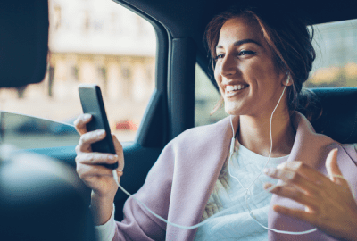Smiling woman wearing earphones holding a smartphone while riding in the back seat of a car