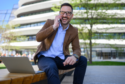 Smiling businessman talking on his phone while sitting outdoors on a bench with a laptop, modern office building in the background