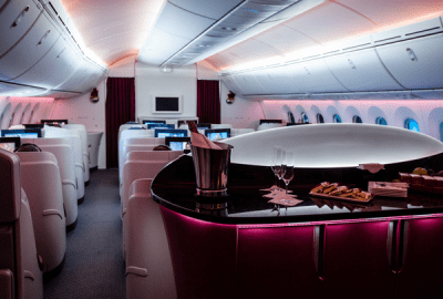 The luxurious interior of a Qatar Airways business class cabin, featuring a curved bar with champagne on ice, wine glasses and snacks, surrounded by private white pod seats with mood lighting
