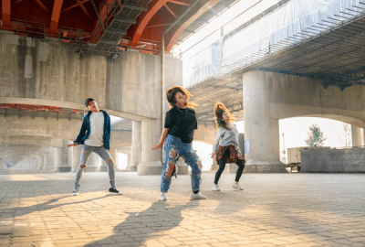 Three young dancers performing K-pop style street dance moves beneath an urban overpass